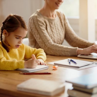 Little girl doing homework while mother is working on a computer at home.