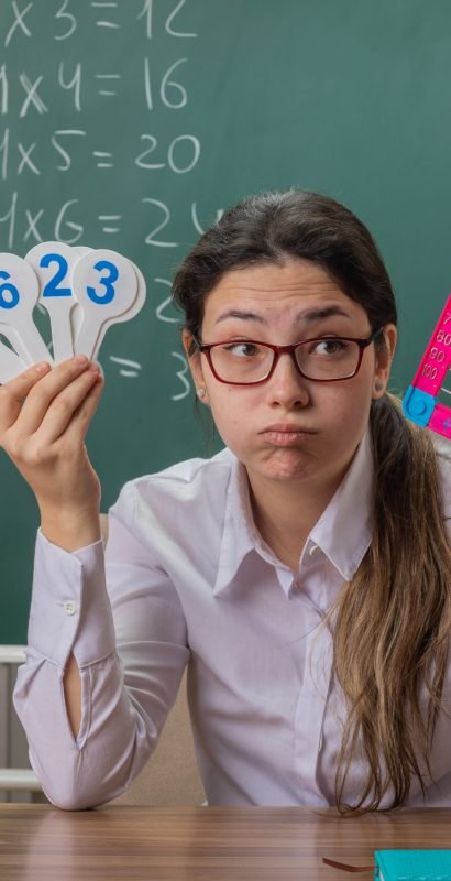 young woman teacher wearing glasses sitting at school desk with bills and number plates looking tired and bored blowing cheeks explaining lesson in front of blackboard in classroom
